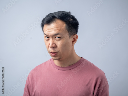 An Asian man in a pink shirt makes a skeptical and uncomfortable expression, as if reacting to something unpleasant. The plain light blue background highlights his displeased