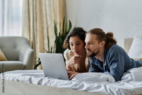 A young couple lies on a bed, looking at a laptop together.