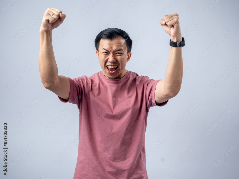 A man in a pink shirt raises both fists in a celebratory gesture, his face beaming with excitement and joy. The plain light blue background emphasizes his triumphant and enthusiastic expression. A man in a pink shirt raises both fists in a celebratory gesture, his face beaming with excitement and joy. The plain light blue background emphasizes his triumphant and enthusiastic expression.