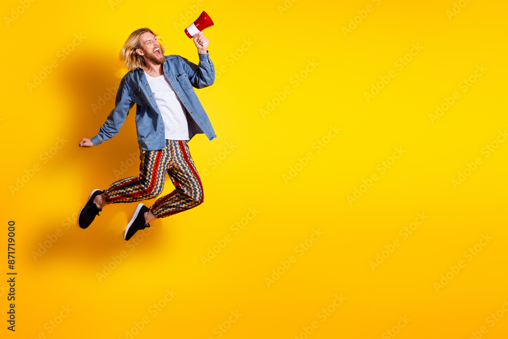 Full size photo of pretty young guy jump scream megaphone wear trendy jeans outfit isolated on yellow color background Full size photo of pretty young guy jump scream megaphone wear trendy jeans outfit isolated on yellow color background