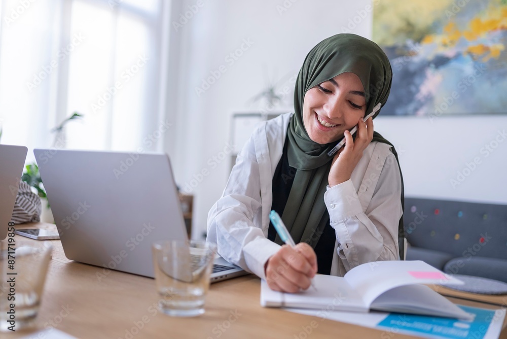 A woman wearing a green hijab, sitting at a desk with a laptop, writing in a notebook while talking on the phone A woman wearing a green hijab, sitting at a desk with a laptop, writing in a notebook while talking on the phone