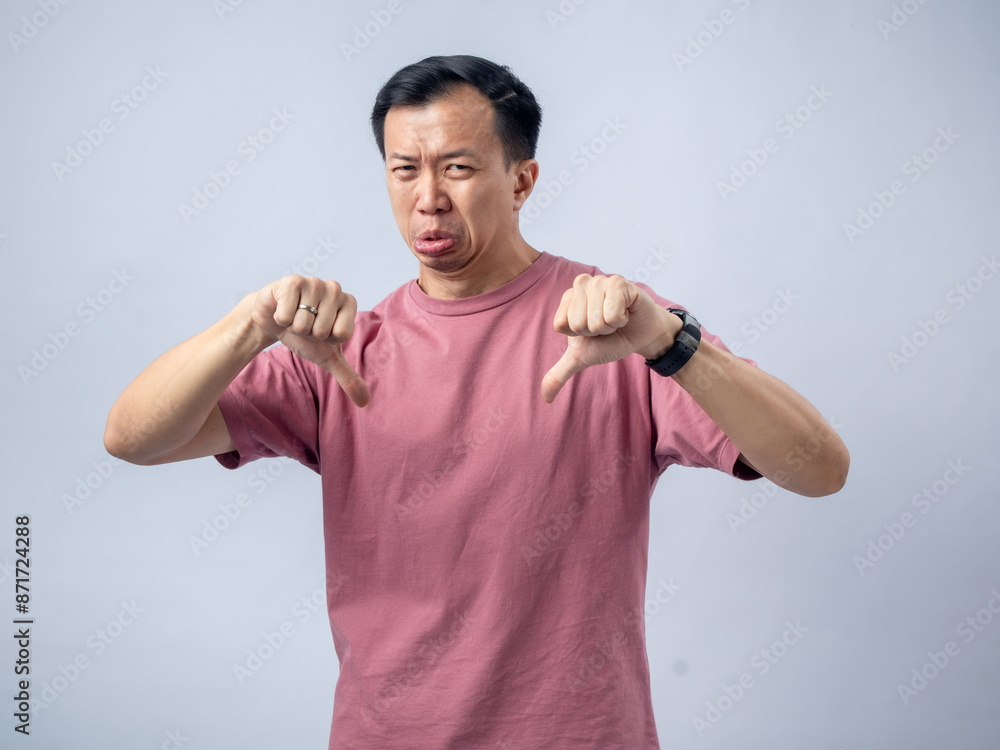 A man in a pink shirt shows a thumbs-down gesture with a displeased expression, sticking out his tongue to indicate disapproval or dissatisfaction. The plain light blue background A man in a pink shirt shows a thumbs-down gesture with a displeased expression, sticking out his tongue to indicate disapproval or dissatisfaction. The plain light blue background