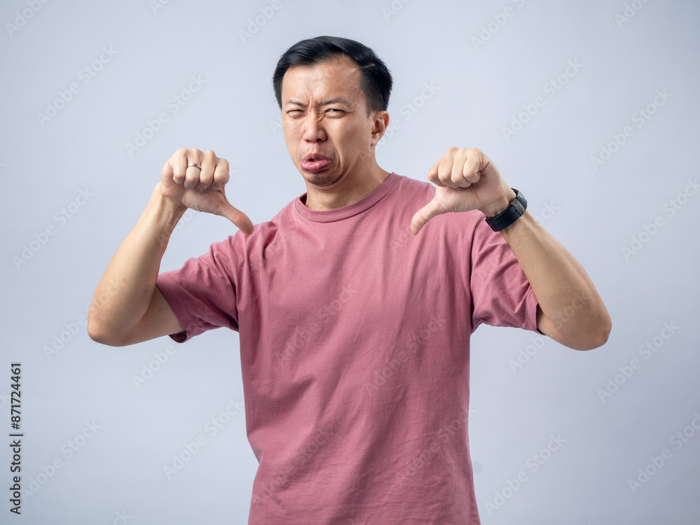 A man in a pink shirt shows a thumbs-down gesture with a displeased expression, sticking out his tongue to indicate disapproval or dissatisfaction. The plain light blue background