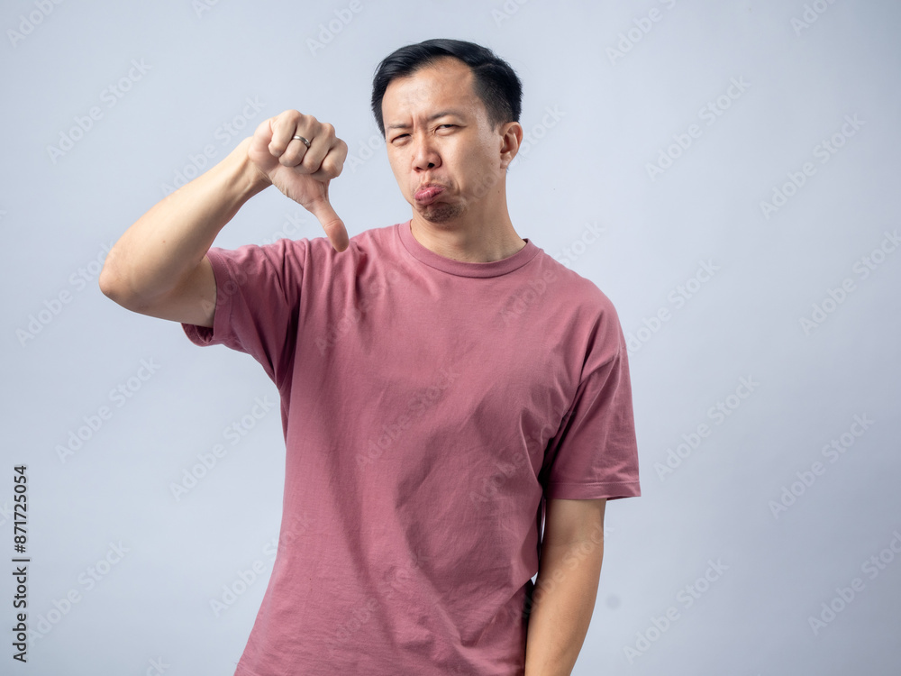 A man in a pink shirt shows a thumbs-down gesture with a displeased expression, sticking out his tongue to indicate disapproval or dissatisfaction. The plain light blue background A man in a pink shirt shows a thumbs-down gesture with a displeased expression, sticking out his tongue to indicate disapproval or dissatisfaction. The plain light blue background