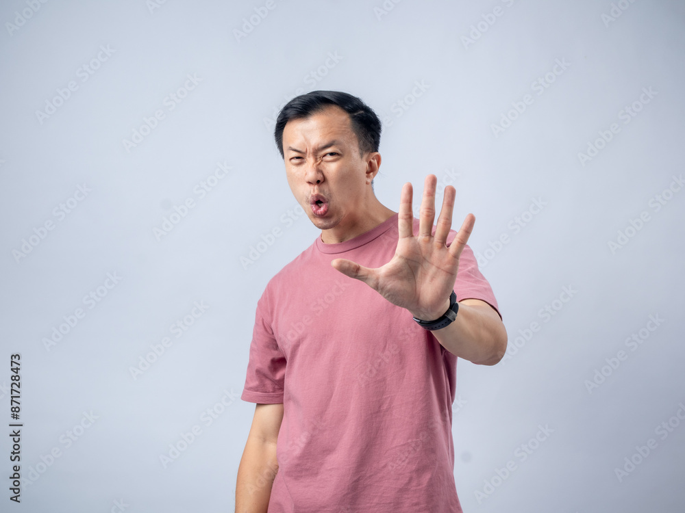 An Asian man in a pink shirt holds up his hand with his palm facing forward, signaling to stop, with a surprised and alert expression. The plain light blue background emphasizes his dramatic An Asian man in a pink shirt holds up his hand with his palm facing forward, signaling to stop, with a surprised and alert expression. The plain light blue background emphasizes his dramatic