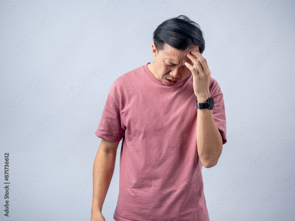 A man in a pink shirt holds his head with a pained expression, indicating a headache or discomfort. The plain light blue background emphasizes his distress and highlights his posture and expression. A man in a pink shirt holds his head with a pained expression, indicating a headache or discomfort. The plain light blue background emphasizes his distress and highlights his posture and expression.