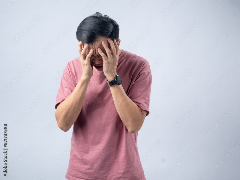A man in a pink shirt holds his head with a pained expression, indicating a headache or discomfort. The plain light blue background emphasizes his distress and highlights his posture and expression. A man in a pink shirt holds his head with a pained expression, indicating a headache or discomfort. The plain light blue background emphasizes his distress and highlights his posture and expression.