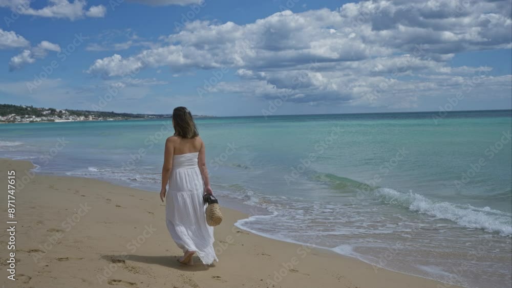 A young hispanic woman in a white dress walks along the sandy beach of pescoluse, salento in puglia, italy, with the turquoise sea and a cloudy sky in the background. A young hispanic woman in a white dress walks along the sandy beach of pescoluse, salento in puglia, italy, with the turquoise sea and a cloudy sky in the background.