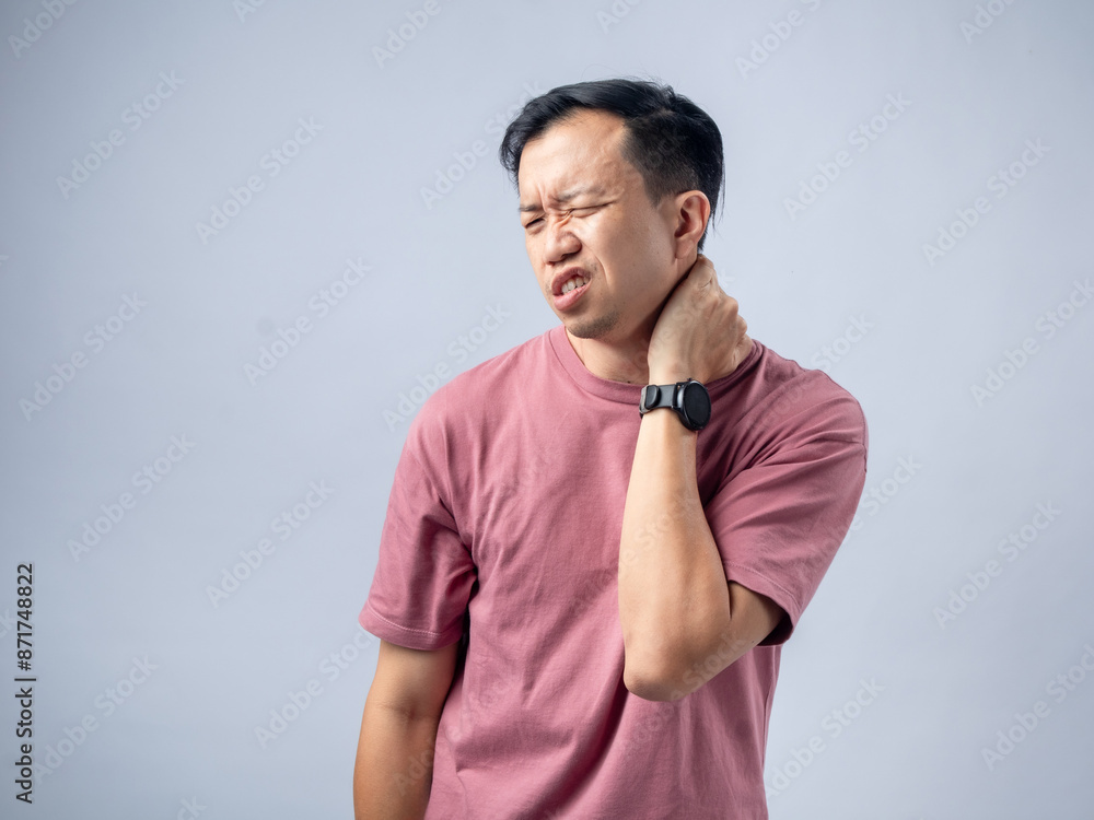 A man in a pink shirt holds his neck with a pained expression, indicating discomfort or neck pain. The plain light blue background emphasizes his distress and highlights A man in a pink shirt holds his neck with a pained expression, indicating discomfort or neck pain. The plain light blue background emphasizes his distress and highlights