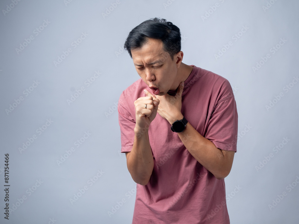 A man in a pink shirt coughs violently, holding his throat with one hand and covering his mouth with the other. His expression shows discomfort and pain. The plain light blue background A man in a pink shirt coughs violently, holding his throat with one hand and covering his mouth with the other. His expression shows discomfort and pain. The plain light blue background