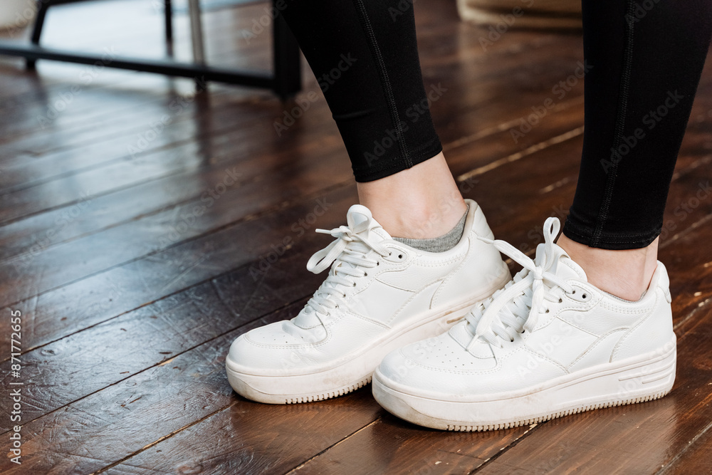 In the scene, there is a person wearing stylish white sneakers, standing on a wooden floor. The footwear contrasts nicely with the natural tones of the wood beneath them In the scene, there is a person wearing stylish white sneakers, standing on a wooden floor. The footwear contrasts nicely with the natural tones of the wood beneath them