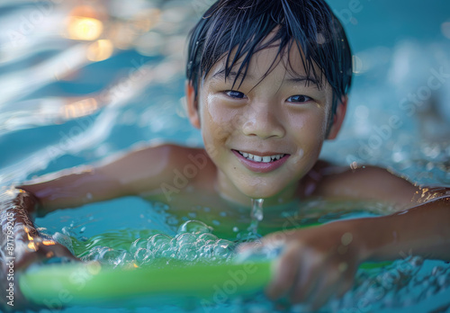 an Asian boy swimming in the pool, wearing goggles and holding a green swim board with his hands. He is smiling happily at the camera