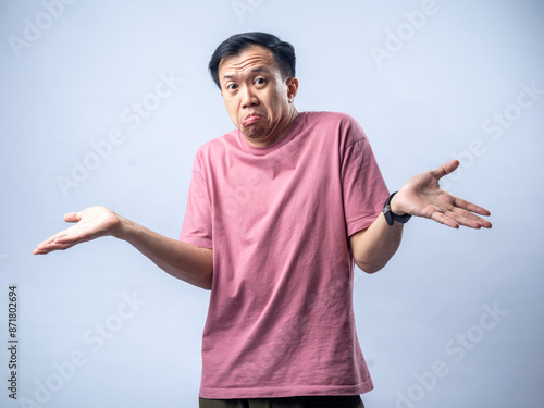 Foto An Asian man in a pink shirt shrugs with a confused and uncertain expression, his arms outstretched and palms up