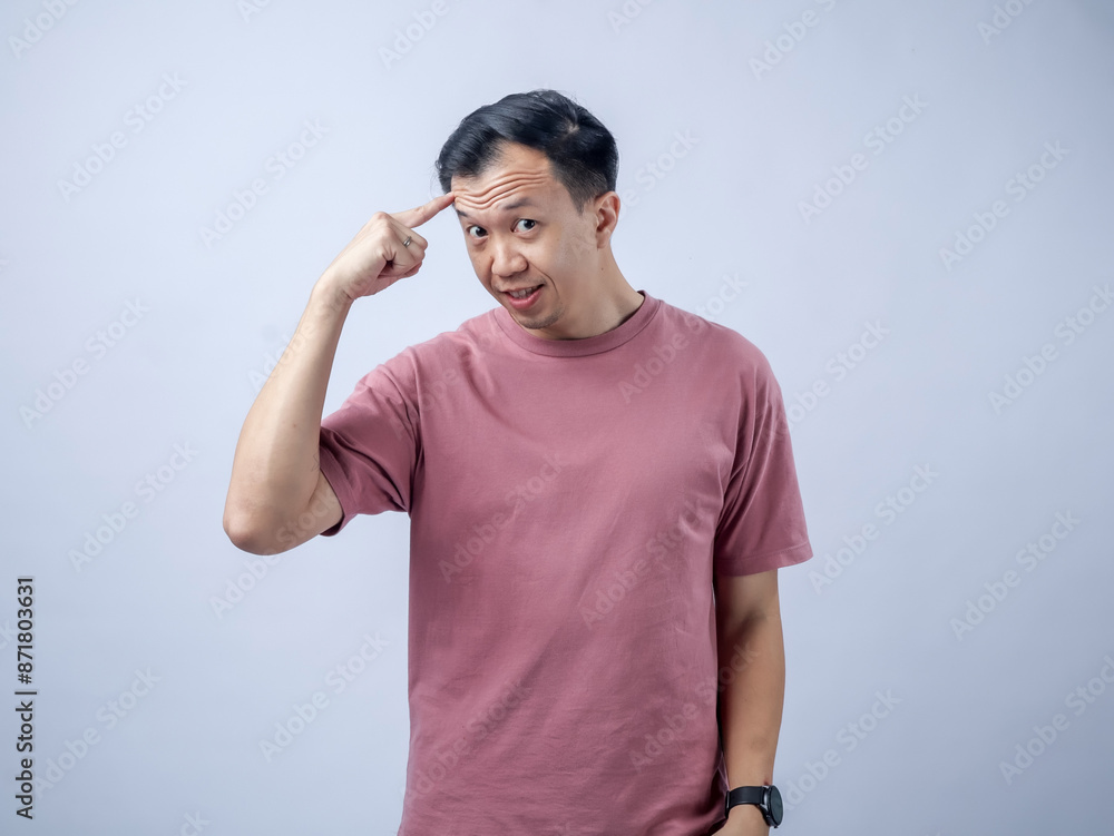 A cheerful Asian man in a pink t-shirt points to his forehead with one finger, smiling confidently. The light grey studio background highlights his casual fashion and thoughtful expression A cheerful Asian man in a pink t-shirt points to his forehead with one finger, smiling confidently. The light grey studio background highlights his casual fashion and thoughtful expression