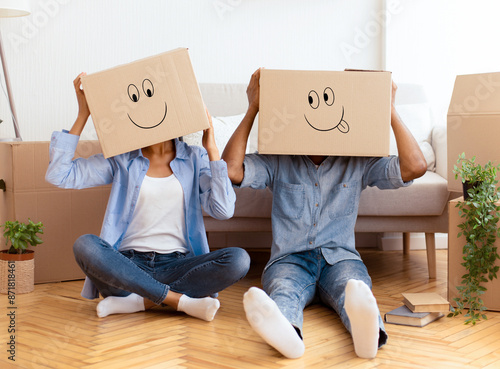 Unrecognizable Afro Couple Wearing Moving Boxes With Smiley Faces On Head Having Fun Sitting On Floor In New Home