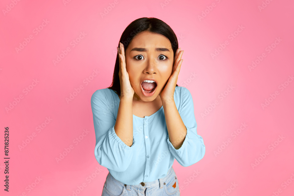 Asian young woman with dark hair wearing a light blue shirt looks shocked with her mouth open and her hands covering her ears. She is standing in front of a pink background. Asian young woman with dark hair wearing a light blue shirt looks shocked with her mouth open and her hands covering her ears. She is standing in front of a pink background.