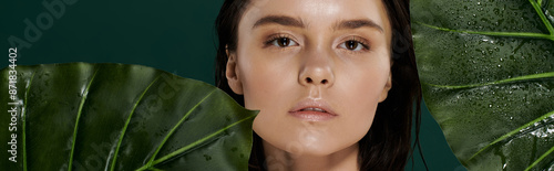A woman with dewy skin gazes softly through lush green leaves.