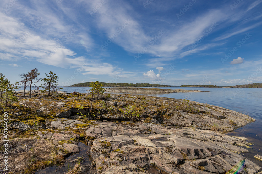 Rocky shoreline with moss and small trees, overlooking a calm, blue lake and distant islands under a bright, clear sky. Rocky shoreline with moss and small trees, overlooking a calm, blue lake and distant islands under a bright, clear sky.