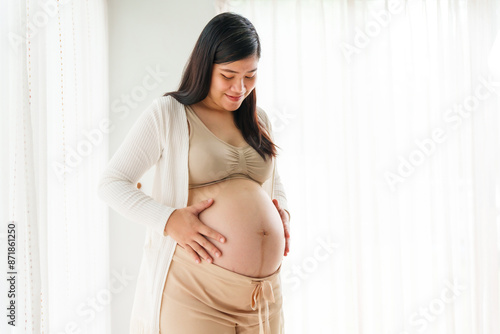 Free Download An Asian young pregnant wife relaxes at home, her hand gently resting on her belly, filled with love and anticipation as she embraces the journey of impending motherhood. - GFXLoad