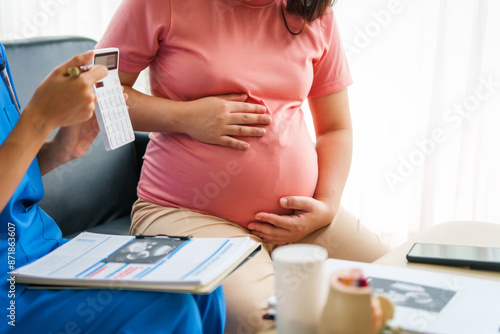 Asian female doctor and nurse consult on a sofa with a pregnant woman, using a calculator to discuss medical expenses, and using ultrasound and stethoscope for thorough examination. photo