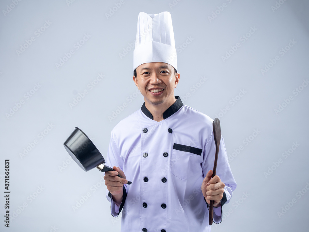 A smiling Asian male chef in a white chef's uniform and hat, holding a pan in one hand and a wooden spoon in the other, conveys friendliness and readiness to cook against a light blue background A smiling Asian male chef in a white chef's uniform and hat, holding a pan in one hand and a wooden spoon in the other, conveys friendliness and readiness to cook against a light blue background