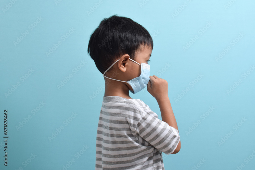 Side portrait of Asian Boy wearing health mask isolated on blue background. Cute little boy holding mask and coughing. Medical and health concept Side portrait of Asian Boy wearing health mask isolated on blue background. Cute little boy holding mask and coughing. Medical and health concept