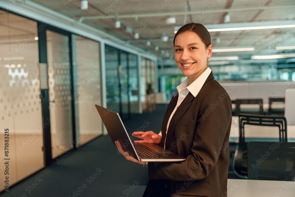 Confident businesswoman is smiling and holding a laptop in a modern open office workspace Confident businesswoman is smiling and holding a laptop in a modern open office workspace
