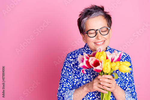 Photo of cheerful person with white gray hairstyle dressed print blouse smell tulips near empty space isolated on pink color background photo