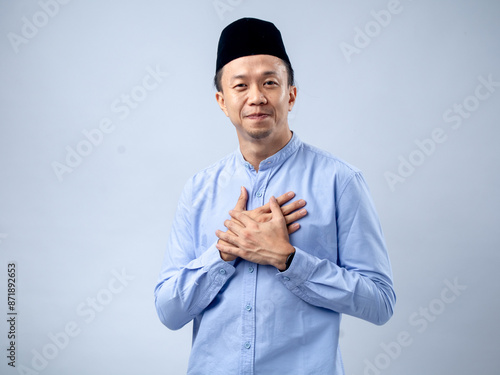 Asian man wearing a black traditional cap and light blue shirt, with hands crossed over his heart, exuding a heartfelt and appreciative expression. The background is plain