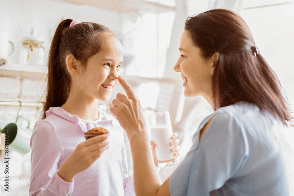 A mother and daughter are enjoying a lighthearted moment in the kitchen. The daughter is holding a cupcake and smiling while the mother playfully touches her nose. A mother and daughter are enjoying a lighthearted moment in the kitchen. The daughter is holding a cupcake and smiling while the mother playfully touches her nose.