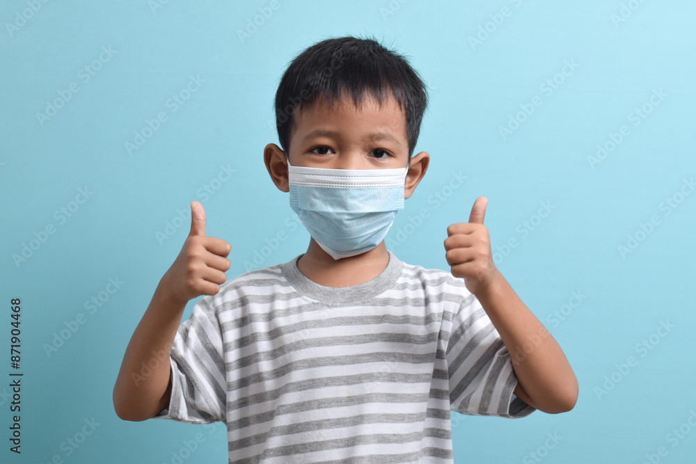 Asian Boy wearing a health mask while giving a thumbs up isolated on a blue background. Medical and health concept. Human population viruses, infections, flu disease