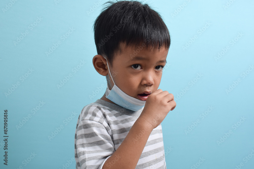 Asian Boy wearing health mask is coughing isolated on blue background. Cute little boy holding mask and coughing. Human population virus, infection, flu disease Asian Boy wearing health mask is coughing isolated on blue background. Cute little boy holding mask and coughing. Human population virus, infection, flu disease