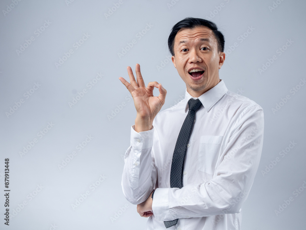 Asian businessman in a white shirt and black tie making an OK gesture with his hand, smiling confidently against a plain light background. The image conveys approval, satisfaction Asian businessman in a white shirt and black tie making an OK gesture with his hand, smiling confidently against a plain light background. The image conveys approval, satisfaction