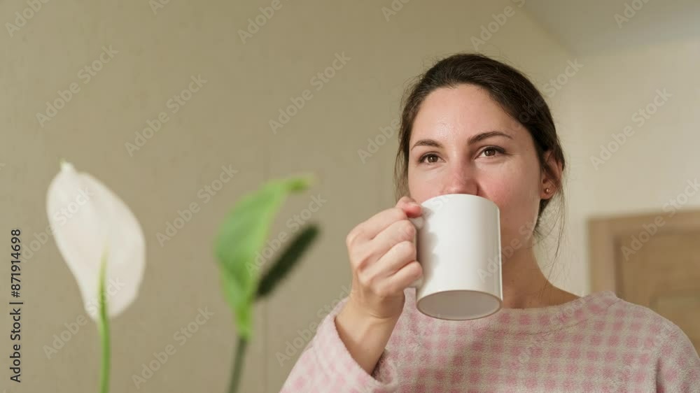 Attractive young adult brunette woman in pajamas takes a sip of coffee from a mug in the kitchen on a sunny morning and looks straight ahead out the window Attractive young adult brunette woman in pajamas takes a sip of coffee from a mug in the kitchen on a sunny morning and looks straight ahead out the window