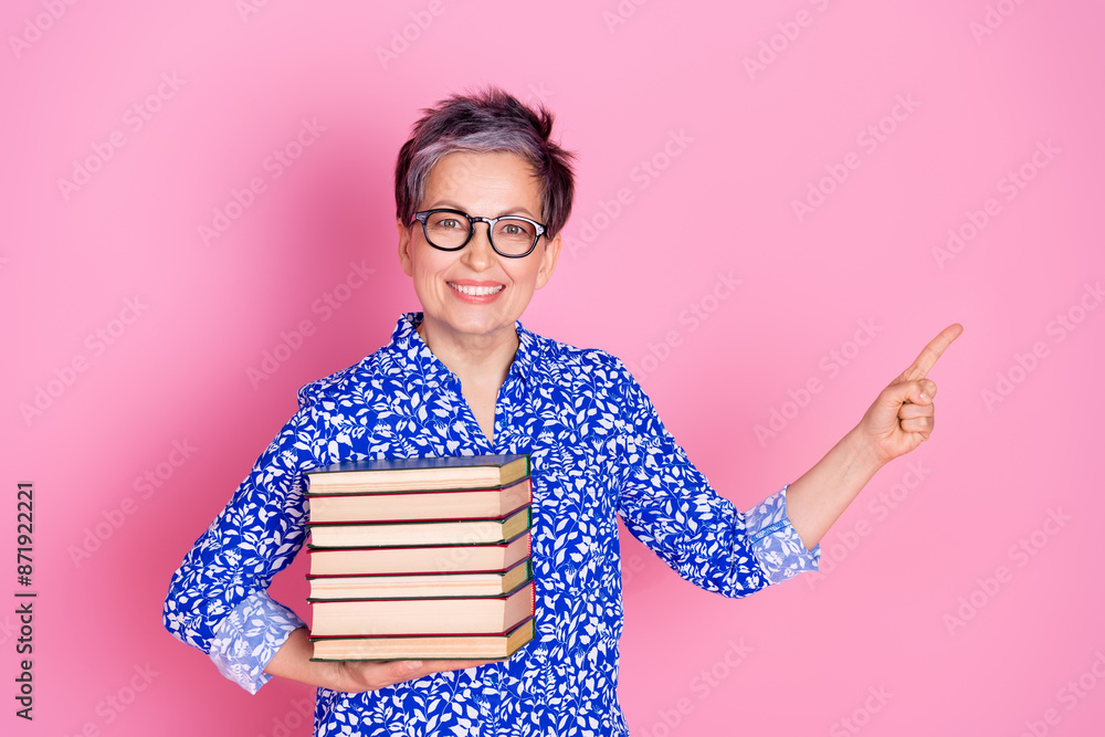 Photo of positive person with gray hair dressed print blouse hold book indicating at offer empty space isolated on pink color background Photo of positive person with gray hair dressed print blouse hold book indicating at offer empty space isolated on pink color background