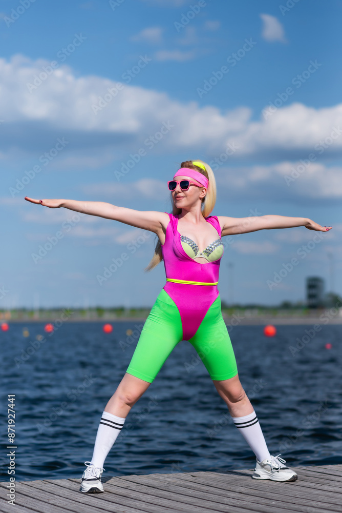 Fototapeta premium Young beautiful blonde woman in sports shorts, a pink bodysuit and pink sunglasses is doing sports outdoors. Doing exercises in sportswear. Sports, Active lifestyle, sports training, healthy lifestyle