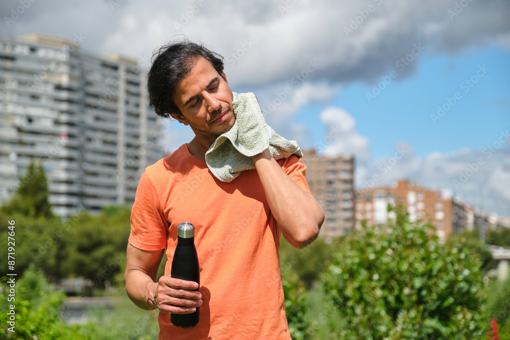 Tired sportsman is wiping sweat with towel and holding bottle of water after workout