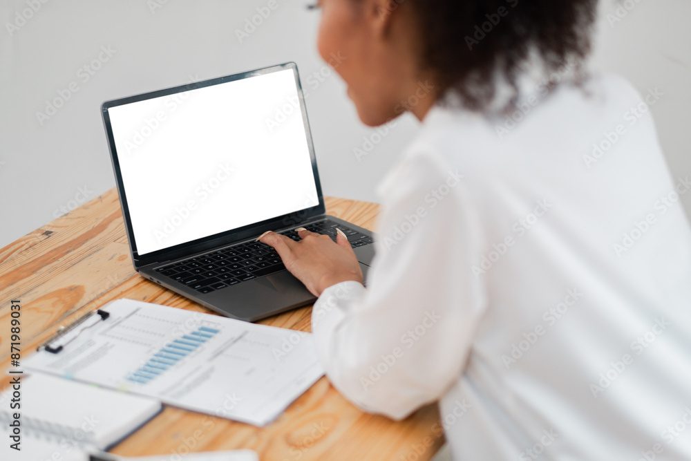Focused woman using laptop at desk with business documents and charts, showcasing a professional work environment. Focused woman using laptop at desk with business documents and charts, showcasing a professional work environment.