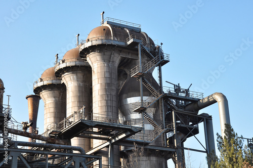 The photograph depicts an old metallurgical plant in Duisburg. Rusty metal structures and pipes dominate the foreground, highlighting the industrial heritage of heavy steel production. The factory's c