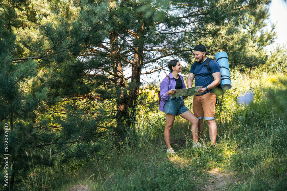 Happy couple tourists with backpacks looking at the map for searching right direction, walking outdoors in the summer forest Happy couple tourists with backpacks looking at the map for searching right direction, walking outdoors in the summer forest