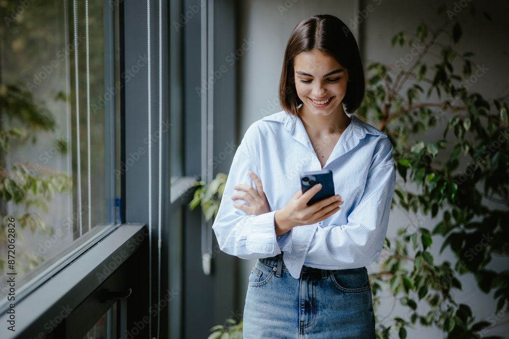 Young woman smiling while using smartphone near window Young woman smiling while using smartphone near window