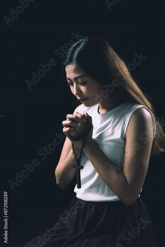 Canvas Print Young woman holding rosary with cross, deep in prayer, against dark background,