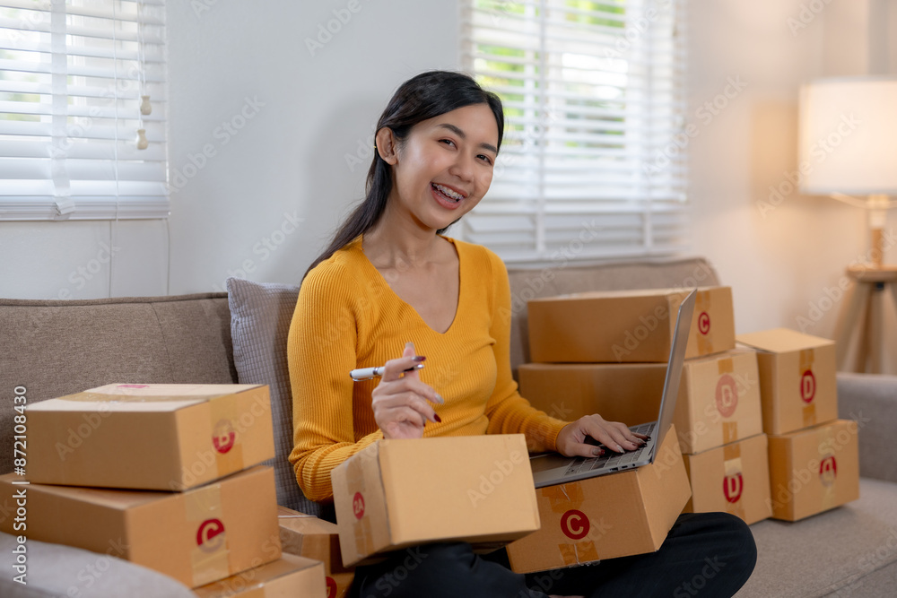 Woman packaging boxes while working on a laptop in a home office environment, representing e-commerce business operations. Woman packaging boxes while working on a laptop in a home office environment, representing e-commerce business operations.