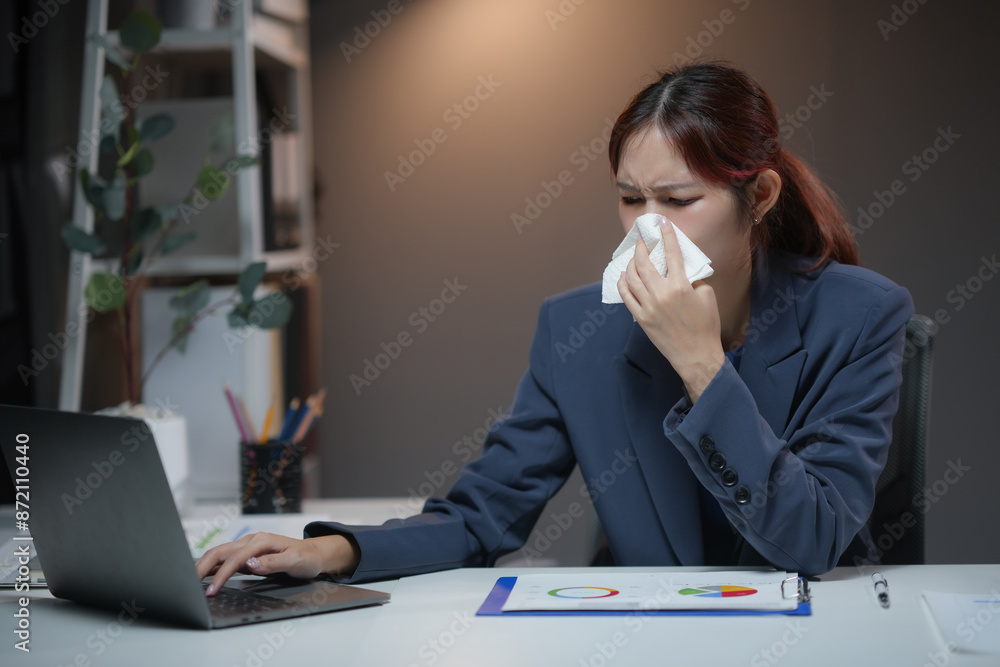 Businesswoman sneezing at the office desk, using a tissue to cover her face with a laptop and documents on the desk during work hours. Businesswoman sneezing at the office desk, using a tissue to cover her face with a laptop and documents on the desk during work hours.