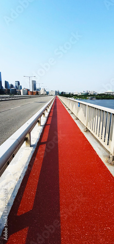 Sogang Bridge on the Hangang River, in Seoul, Korea, with a pedestrian path with a red surface and a road for vehicles, various high-rise buildings, indicating a cityscape. The sky is clear and blue photo