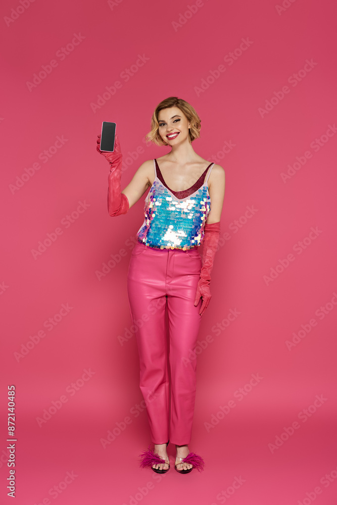 A woman in pink pants and a sequined top holds a smartphone against a pink background. A woman in pink pants and a sequined top holds a smartphone against a pink background.