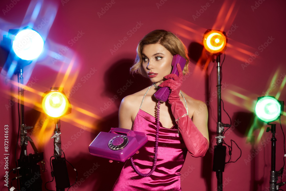 A woman in a pink dress and gloves speaks on a vintage phone while posing under studio lights. A woman in a pink dress and gloves speaks on a vintage phone while posing under studio lights.