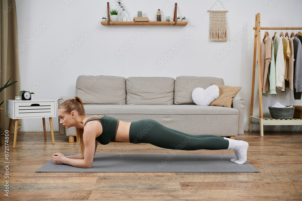 Fototapeta premium A young woman in athletic wear performs a plank exercise on a yoga mat in her living room.