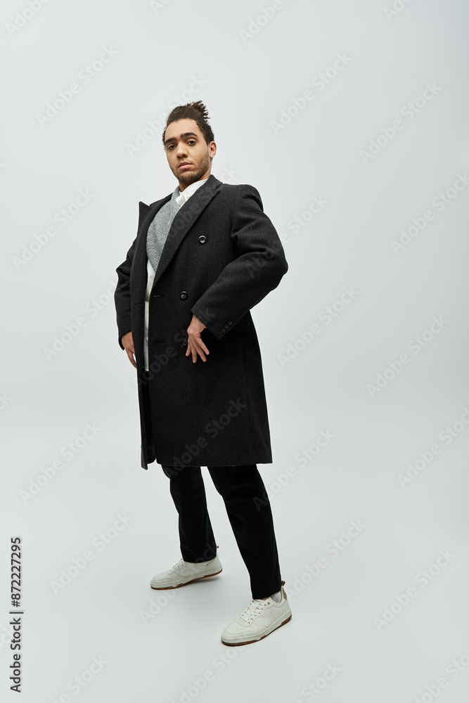 A stylish African American man stands confidently in a black coat and white sneakers against a grey background. A stylish African American man stands confidently in a black coat and white sneakers against a grey background.