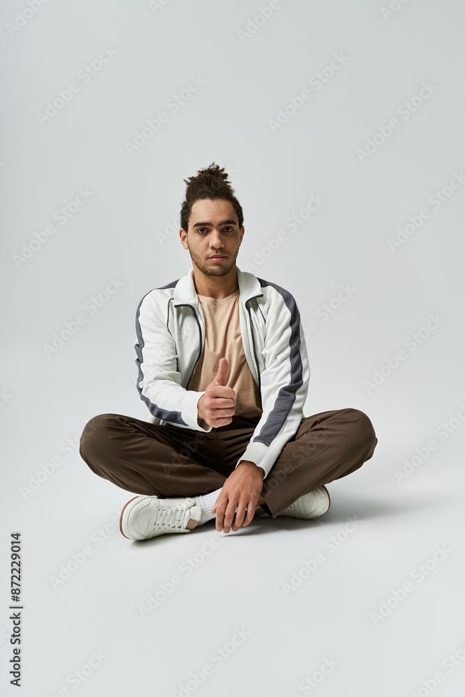A young African American man sits with his legs crossed, wearing a white jacket and brown pants, against a gray background. A young African American man sits with his legs crossed, wearing a white jacket and brown pants, against a gray background.